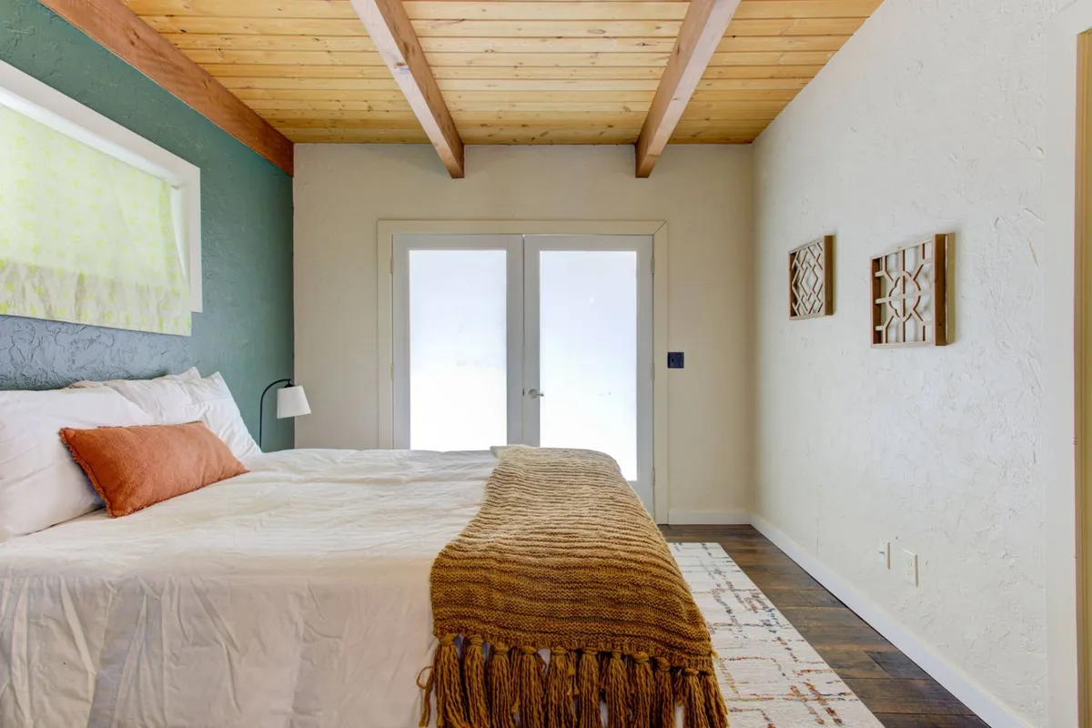 A tranquil bedroom featuring a bed with white linens and an orange accent pillow, wooden ceiling beams, and glass doors that open to outside views. The space exudes comfort and quiet, ideal for relaxation and enjoying morning coffee.