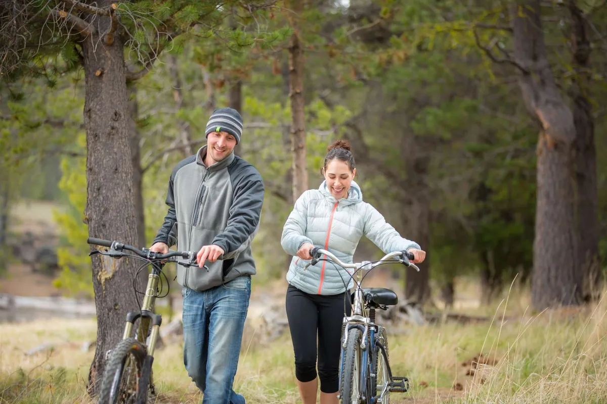Two individuals walk their bicycles along a forested path, surrounded by lush greenery and natural light. The scene suggests warm-weather adventures such as hiking and biking, complementing the winter-focused images.