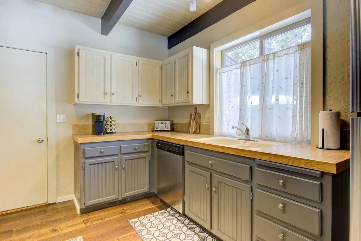 A cozy kitchen corner displaying cabinetry, countertop space, a window with a curtain, a sink, dishwasher, and a coffee maker. The space feels intimate and functional, inviting shared breakfasts or casual meal prep.