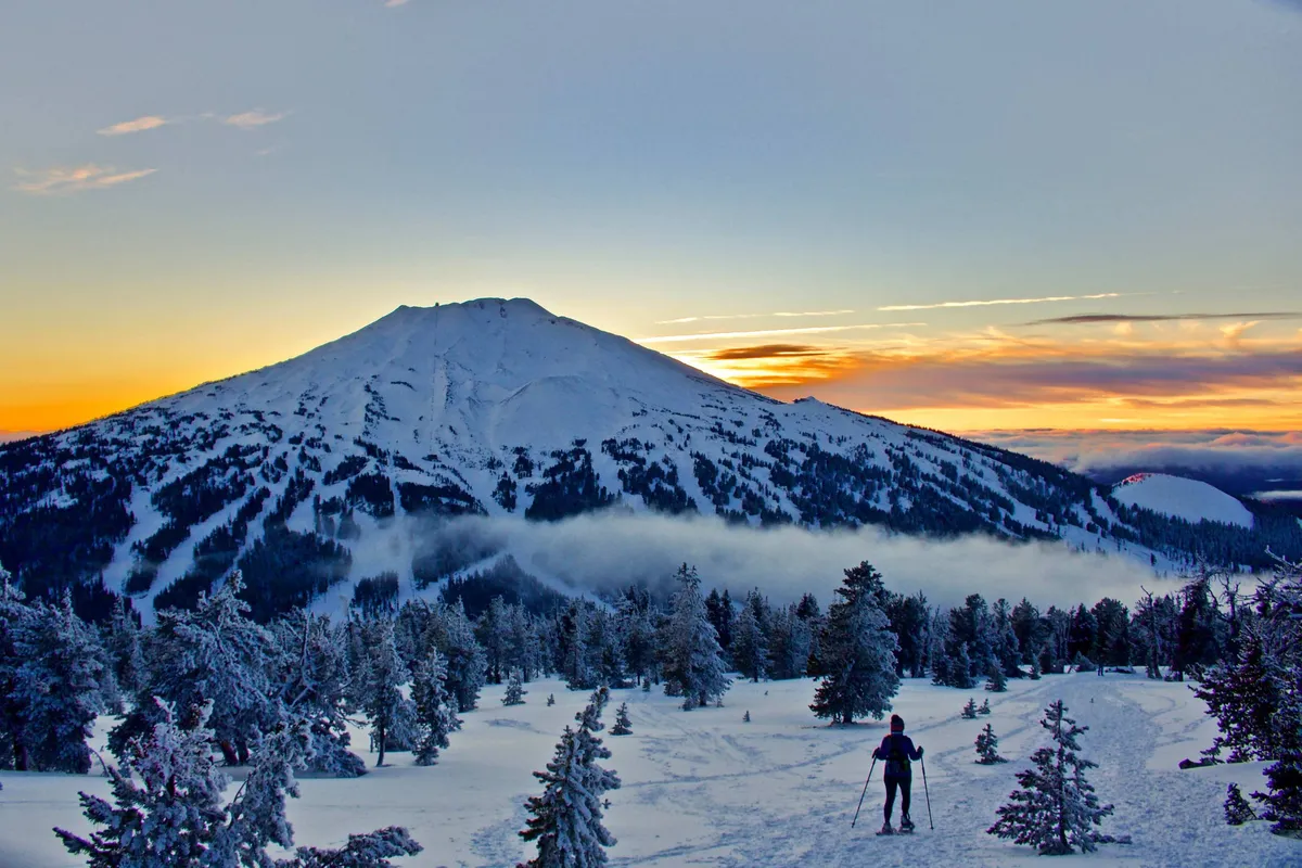 A breathtaking panoramic view of a snow-covered mountain at sunset, with a lone skier silhouetted against the colorful sky. The landscape is dramatic, highlighting both the adventure and tranquility of mountain activities.