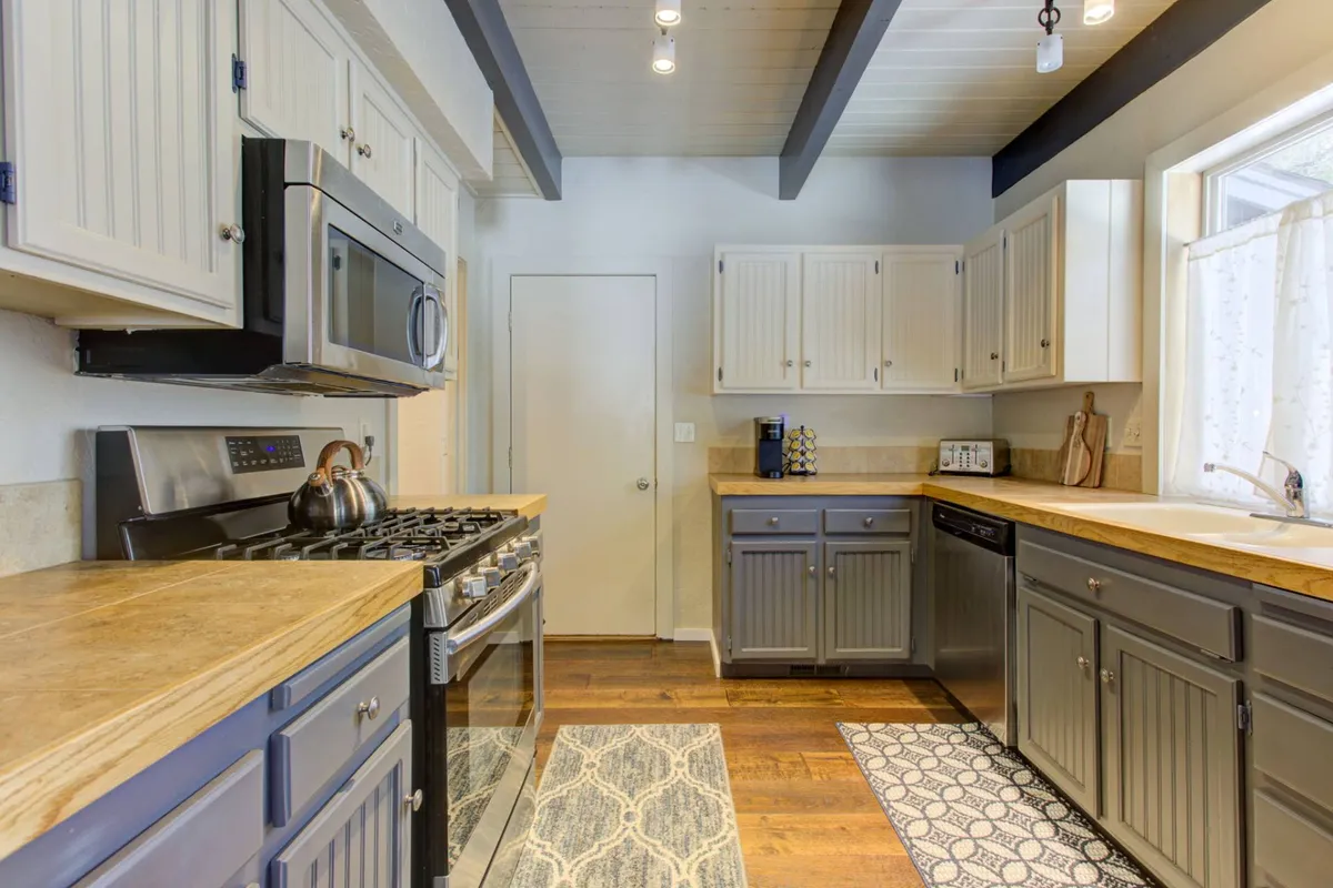 This image features a kitchen space with stainless steel appliances, warm wooden countertops, a decorative rug, and a window letting in abundant natural light. The scene is inviting and visually demonstrates the kitchen as a central, open area ideal for cooking and socializing.