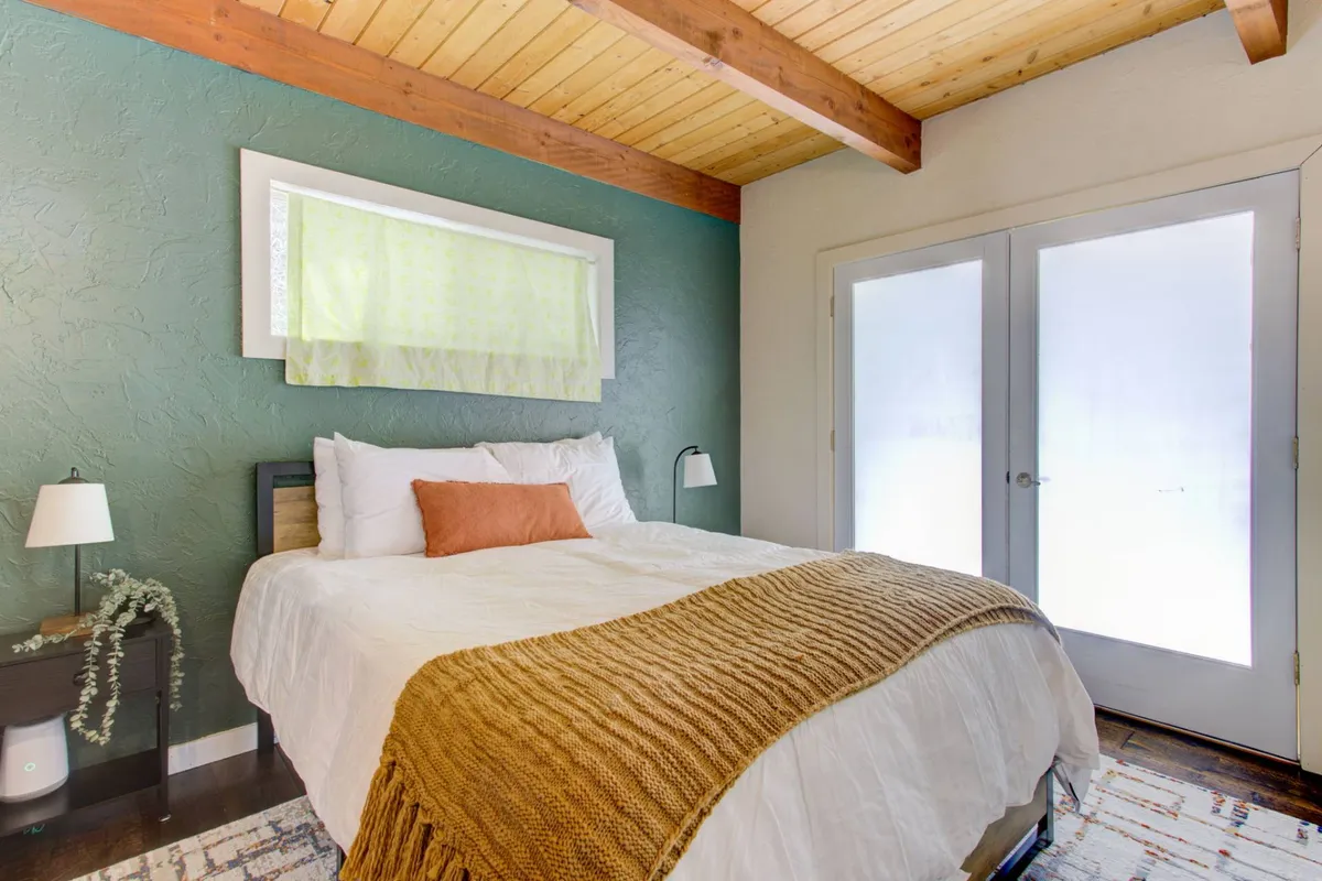 A cozy bedroom featuring a bed with crisp white bedding and an orange accent pillow. The room is accented by green walls, exposed wooden ceiling beams, and large glass doors that let in abundant natural light and offer tranquil views of the forest beyond.