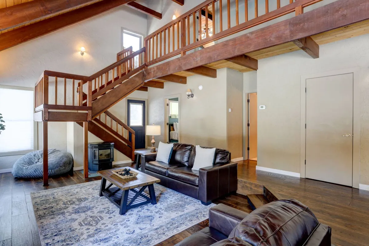 A warm, inviting living space featuring leather sofas, a decorative rug, and a striking wooden staircase. Large windows frame views of the surrounding forest, filling the room with natural light. The ambiance is cozy and suited for relaxation, reading, or remote work by the fire.