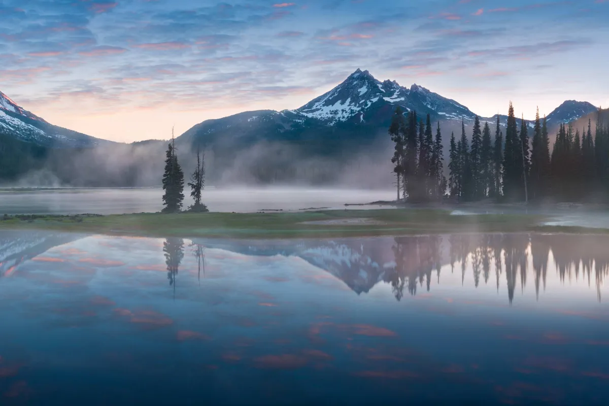 A breathtaking view of distant mountains, a peaceful lake, and surrounding trees under an expansive sky. The scene suggests opportunities for hiking, birdwatching, or simply soaking in the region's natural splendor.
