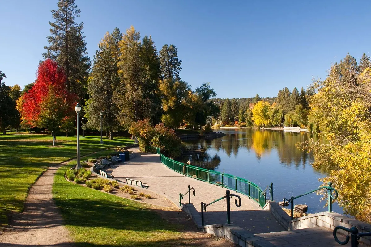 A tranquil riverside scene with a stone pathway, colorful autumn trees, calm water, and park benches, inviting visitors to stroll, picnic, or simply enjoy the peaceful environment. The image conveys a sense of community and seasonal transformation.