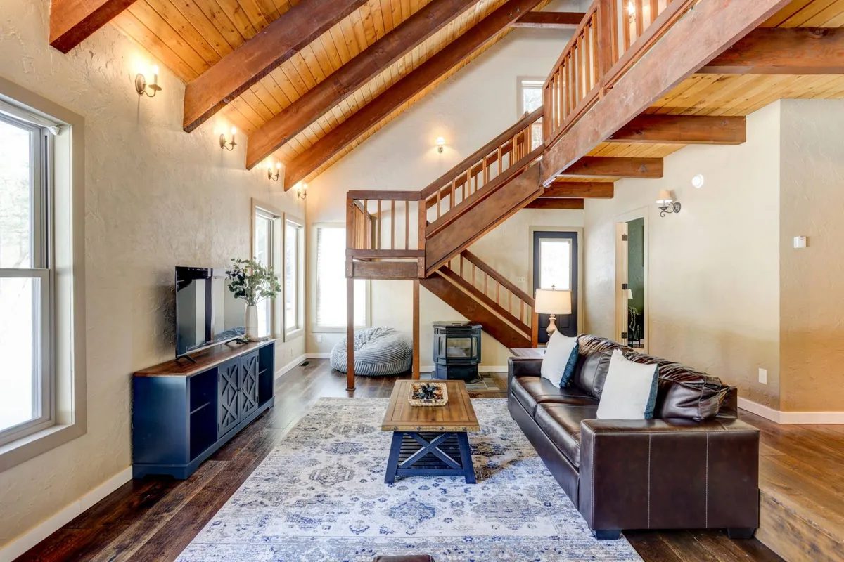 The image showcases a warm, inviting living room with a plush sofa, a stylish coffee table, a modern staircase, a large television, and expansive windows that frame breathtaking views of the surrounding pines. The open-concept space is ideal for relaxing, gathering with loved ones, and enjoying cozy evenings together.