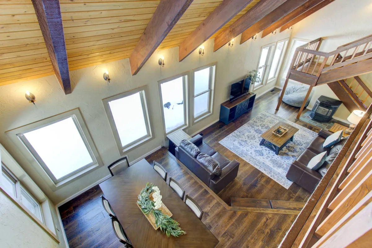 This image captures a spacious, open-concept living and dining area. The focal points include a large dining table capable of seating a group, a plush sofa area, expansive windows that frame views of tall pine trees, and architectural wooden beams that add warmth and character to the space. The arrangement suggests a welcoming environment ideal for gatherings, shared meals, and time spent together.