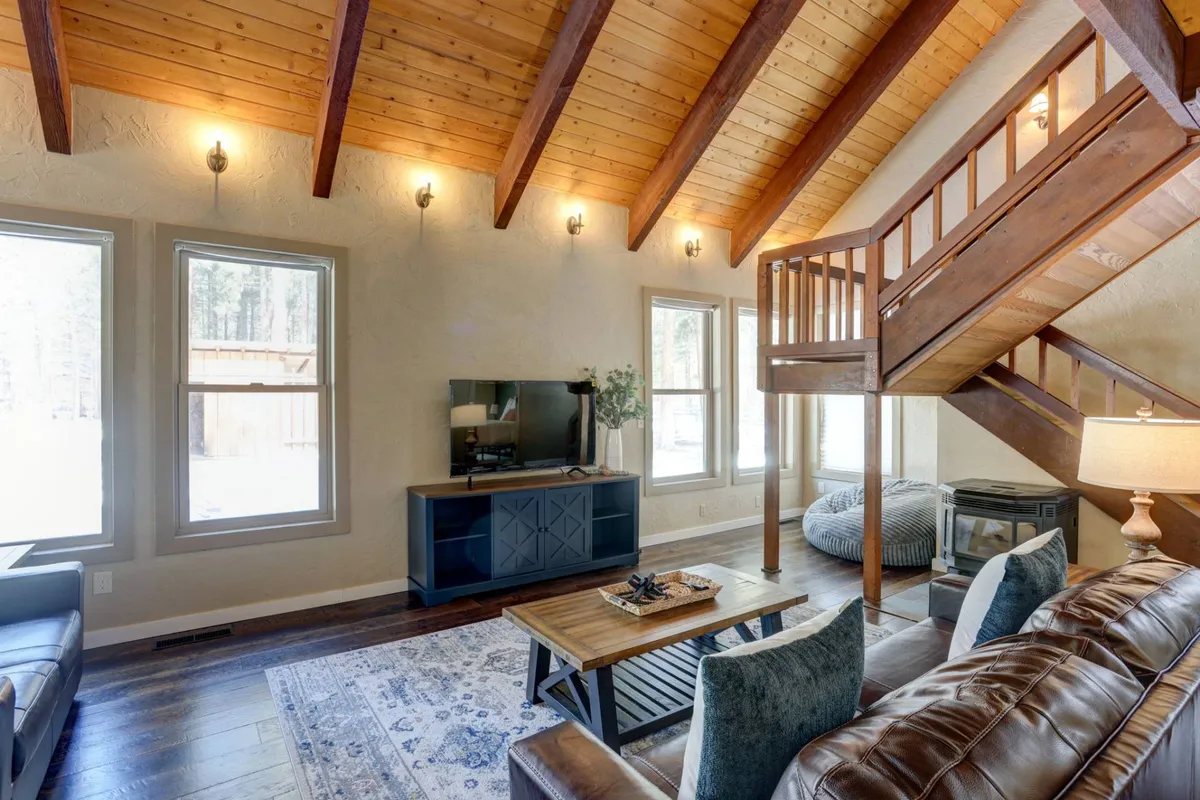 The living room is pictured here with a plush sofa, a central coffee table perfect for games or casual conversations, a staircase accent, a flat-screen TV for streaming, and wide windows providing ample natural light and views of the surrounding pines.