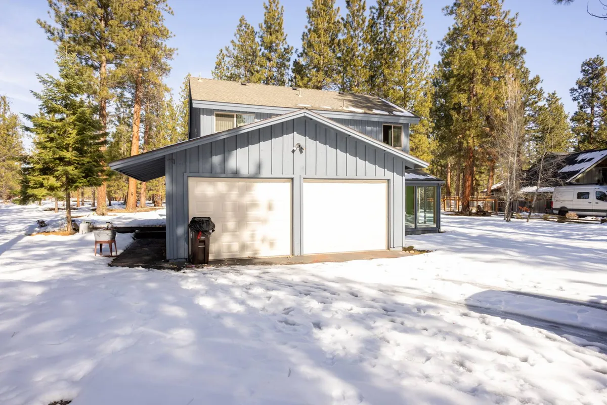 A peaceful scene featuring a two-car garage, snow-covered ground, and tall pine trees that frame the blue-siding house, presenting a sense of tranquility and room to roam.